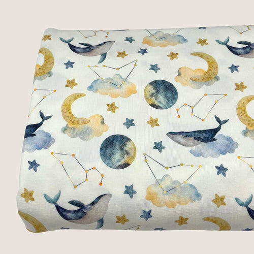 A piece of **Cotton Jersey Fabric - Moon Whales** by **Once Upon A Fabric**, featuring a whimsical design with whales, moons, stars, clouds, and constellations in soft pastel colors. Perfect for creating adorable baby clothes, the background is white with illustrations in shades of blue, yellow, and gray. Oeko-Tex 100 certified.