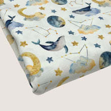 Load image into Gallery viewer, A piece of **Once Upon A Fabric Cotton Jersey Fabric - Moon Whales** with a whimsical pattern featuring whales, stars, moons, clouds, and constellations in various shades of blue, yellow, and gray. Oeko-Tex 100 certified and perfect for baby clothes, the design is set against a white background.
