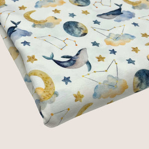 A piece of **Once Upon A Fabric Cotton Jersey Fabric - Moon Whales** with a whimsical pattern featuring whales, stars, moons, clouds, and constellations in various shades of blue, yellow, and gray. Oeko-Tex 100 certified and perfect for baby clothes, the design is set against a white background.