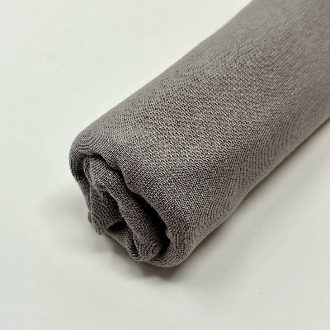 A rolled-up gray fabric placed on a white surface. The fabric, MiskaHryzka Organic Tubular Cuff Ribbing - MEDIUM GREY, made from Organic GOTS certified material, appears smooth and evenly rolled with a slightly textured surface.