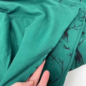 A hand is holding up a piece of LAST METER French Terry Fabric - Woodland Dark Green from Once Upon A Fabric with black deer and tree branch patterns. The person appears to be examining the edge seam. The background is out of focus, showing more Woodland print fabric with the same pattern, all Oeko-Tex 100 certified.