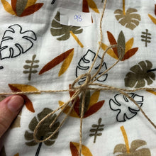 Load image into Gallery viewer, A piece of REMNANT 98 CM Double Gauze - Autumn Leaves from Once Upon A Fabric with a pattern of orange, brown, and beige leaves, and some black and white outline leaves. The fabric is tied with a piece of twine, and a small paper with the number &quot;98&quot; is placed on it. A hand is holding this perfect addition to baby items.
