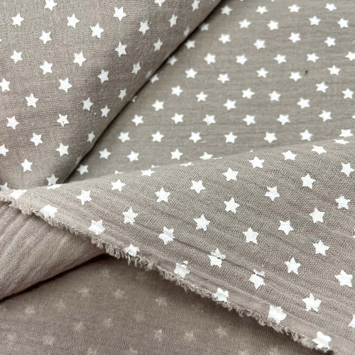 A close-up image of Muslin Fabric / Double Gauze Fabric - Mini Stars Taupe by Once Upon A Fabric with a grayish-brown background featuring a pattern of small white stars. The Muslin Fabric / Double Gauze Fabric - Mini Stars Taupe is layered, with the edges slightly frayed.