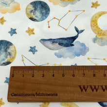 Load image into Gallery viewer, A wooden ruler marked in centimeters and millimeters is placed on white Cotton Jersey Fabric - Moon Whales from Once Upon A Fabric with printed designs of moons, stars, constellations, and a whale resting on a cloud, perfect for creating Oeko-Tex 100 certified baby clothes.
