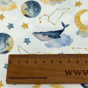 A wooden ruler marked in centimeters and millimeters is placed on white Cotton Jersey Fabric - Moon Whales from Once Upon A Fabric with printed designs of moons, stars, constellations, and a whale resting on a cloud, perfect for creating Oeko-Tex 100 certified baby clothes.