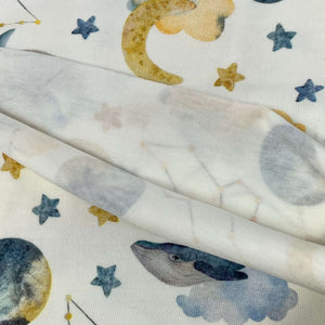 A close-up image of fabric with a celestial-themed design, perfect for baby clothes. The pattern includes images of moons, stars, and a whale, all in muted colors. Made from soft *Once Upon A Fabric* Cotton Jersey Fabric - Moon Whales*, Oeko-Tex 100 certified for safety and quality, it appears partly folded.