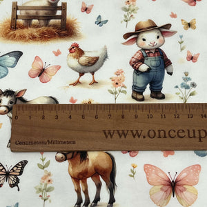 A whimsical farm-themed organic cotton jersey fabric features a farm, a lamb in a hay-filled pen, butterflies, flowers, a chicken, a donkey, and a horse, all set against a light background. Organic GOTS Cotton Jersey - Farm by Once Upon A Fabric.