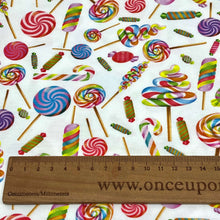 Load image into Gallery viewer, A close-up of a fabric pattern featuring various colorful candy designs such as lollipops and wrapped candies on a white background. Made from GOTS certified organic cotton, this Organic GOTS Cotton Jersey - Lollipops by Once Upon A Fabric also shows a wooden ruler at the bottom marked in centimeters and millimeters, with &quot;www.onceupon&quot; partially visible.
