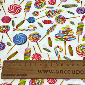 A close-up of a fabric pattern featuring various colorful candy designs such as lollipops and wrapped candies on a white background. Made from GOTS certified organic cotton, this Organic GOTS Cotton Jersey - Lollipops by Once Upon A Fabric also shows a wooden ruler at the bottom marked in centimeters and millimeters, with "www.onceupon" partially visible.