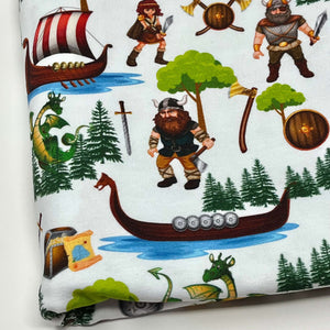 Organic cotton jersey fabric featuring Vikings, longships, dragons, treasure chests, axes, pine trees, and shields. The elements are scattered across a white background with some Vikings holding weapons and two green dragons depicted.