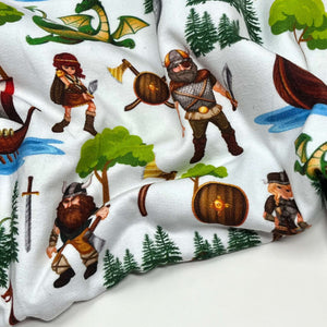 Organic cotton jersey fabric featuring Vikings, longships, dragons, treasure chests, axes, pine trees, and shields. The elements are scattered across a white background with some Vikings holding weapons and two green dragons depicted.