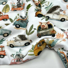 Load image into Gallery viewer, Organic GOTS Cotton Jersey with a whimsical pattern featuring small animals driving colourful cars. The digitally printed design includes mice, rabbits, hedgehogs, and elephants, surrounded by various plants and flowers against a white background.

