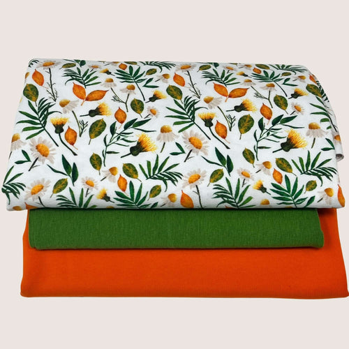 A stack of three folded cotton jersey fabrics. The top fabric is white with an orange, yellow, and green floral pattern. The middle fabric is solid green, and the bottom fabric is solid orange. All fabrics are neatly folded and stacked on top of each other. This is the Jersey Remnants Bundle 1.3 m from Once Upon A Fabric.