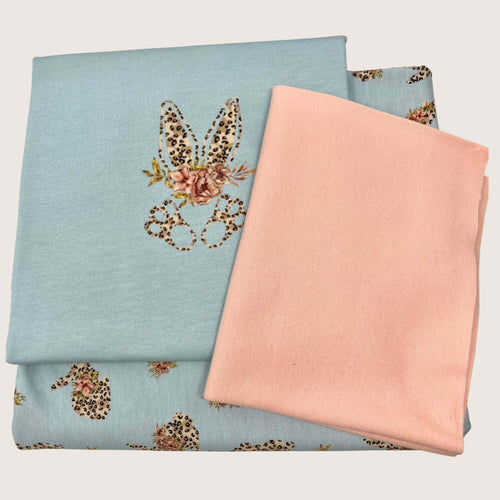 The image shows folded cotton jersey fabrics in two different designs. One fabric is light blue with a leopard print bunny and floral design, while the other is solid pink. The Jersey Remnants Bundle 1.7 m by Once Upon A Fabric, perfect for jersey panels, are arranged in a neat stack.
