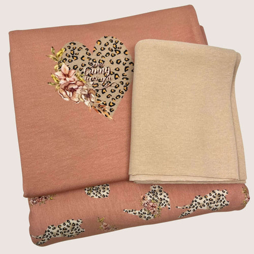 Two folded clothing items placed on a light background. One is a pink fabric with a leopard print heart and floral design, crafted from cotton jersey fabrics. The other is a plain beige fabric, both appearing soft and suitable for apparel or accessories. These fabrics are part of the Jersey Remnants Bundle 1.9 m by Once Upon A Fabric.