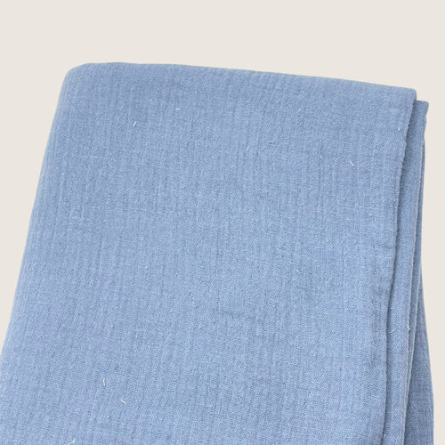 A folded piece of Muslin Fabric / Double Gauze Fabric - Light Vintage Blue from Once Upon A Fabric with a textured pattern. The material appears to be a soft, woven gauze fabric, neatly arranged. The background is plain and off-white.