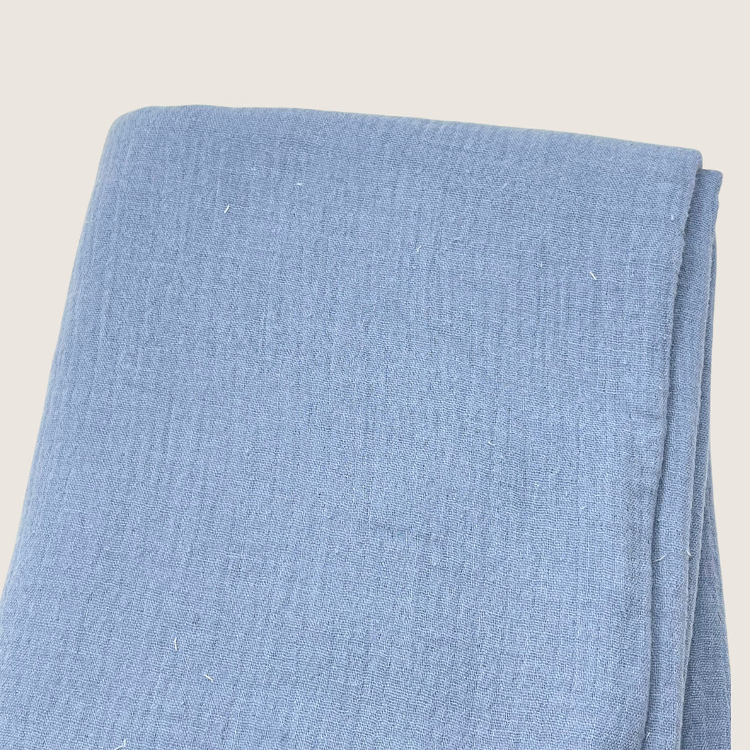 A folded piece of Muslin Fabric / Double Gauze Fabric - Light Vintage Blue from Once Upon A Fabric with a textured pattern. The material appears to be a soft, woven gauze fabric, neatly arranged. The background is plain and off-white.