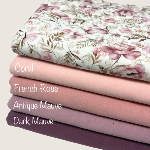 A stack of five folded cotton jersey fabrics arranged in a gradient from light to dark is displayed. The top fabric, perfect for baby clothes, has a floral pattern with shades of pink and green. The Oeko-Tex 100 certified fabrics are labeled from top to bottom: Solid Cotton Jersey Fabric - French Rose by Once Upon A Fabric, Coral, Antique Mauve, and Dark Mauve.