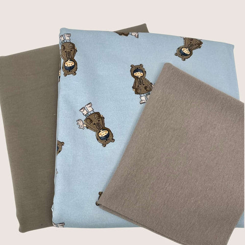 Three pieces of fabric are shown. One piece, a light blue French terry fabric, has illustrations of a person wearing a coat, boots, and a hat. The other two pieces are plain cotton jersey fabrics, with one being dark gray and the other light gray. The fabrics are neatly arranged in a stack. This is the Jersey and French Terry Remnants Bundle 1.5 m from Once Upon A Fabric.
