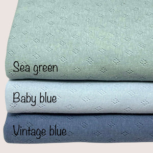 Three folded fabrics with labels are stacked vertically. From top to bottom: sea green, baby blue, and Pointelle Jersey - Vintage Blue by Once Upon A Fabric. Each lightweight fabric displays a subtle diamond pattern and is crafted from pointelle cotton jersey, perfect for creating soft baby clothes.