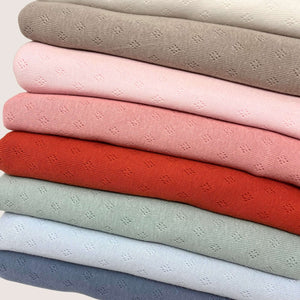 A stack of folded Pointelle Jersey - French Rose fabrics by Once Upon A Fabric in various colors. From top to bottom, the colors are beige, light pink, darker pink, coral, red, light green, light blue, and blue. Each lightweight cotton fabric has a small, repeating eyelet pattern and is Oeko-Tex 100 certified.