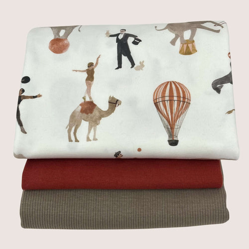 Three folded fabrics are stacked on top of each other. The top fabric, a rib knit jersey, features an illustrated circus theme with performers, animals, and a hot air balloon on a white background. The middle fabric is solid red cotton jersey, and the bottom fabric is solid gray. This trio is part of the 