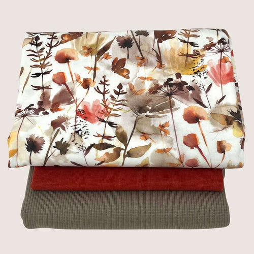 A stack of three folded fabrics is shown. The top fabric, an organic GOTS cotton jersey, features a floral print with autumn colors. The middle one is a solid burnt orange rib knit jersey, and the bottom one is a solid brownish-gray color. The Jersey Remnants Bundle 1.3 m from Once Upon A Fabric is displayed against a plain background.
