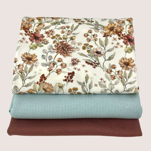 Three folded blankets are stacked. The top blanket, crafted from cotton jersey, features a floral pattern with green leaves and various flowers on a white background. The middle blanket is solid light teal, and the bottom blanket is solid brown. The background is off-white. This collection of fabrics is part of the Jersey Remnants Bundle 1.25 m by Once Upon A Fabric.