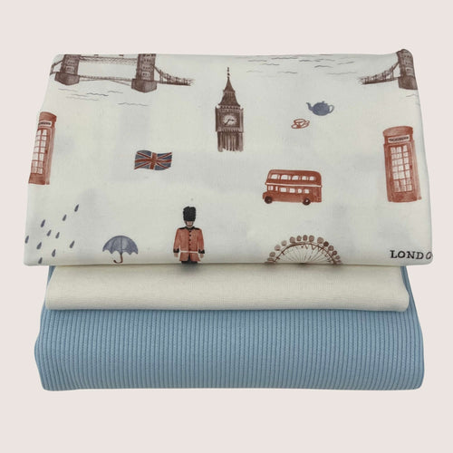 Three folded fabric pieces are stacked, with the top piece showcasing London-themed illustrations like the Tower Bridge, Big Ben, a red double-decker bus, a British guard, and a Union Jack flag. The middle piece is cream-colored cotton jersey fabric, and the bottom piece is light blue. This is the Jersey Remnants Bundle 1 m from Once Upon A Fabric.