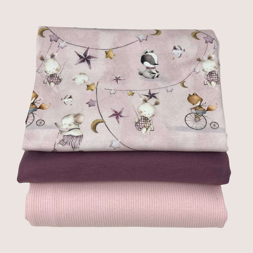 Three folded Jersey Remnants Bundle 1.4 m from Once Upon A Fabric are stacked on top of each other. The top fabric has a design featuring whimsical animals, stars, and moons on a light pink background. The middle fabric is solid purple cotton jersey, and the bottom fabric is light pink with a ribbed texture.