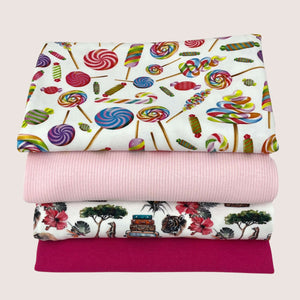Four folded fabrics are stacked on top of each other. The top fabric features a colorful candy print, the second is a solid pink, the third has a scenic print with trees and vehicles, and the bottom fabric is in solid dark pink. Made entirely from organic cotton, these fabrics promise both comfort and quality. This Jersey Remnants Bundle 1.5 m by Once Upon A Fabric is perfect for your next sewing project.