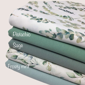 A stack of four folded baby clothes fabrics reveals their colors and patterns. The top cotton jersey fabric, labeled "Solid Cotton Jersey Fabric - Pistachio" by Once Upon A Fabric, features leaf prints, the second, "Sage," is a solid color, the third "Frosty Mint" displays leaf prints, and the bottom in a solid blue-green hue. All are Oeko-Tex 100 certified.