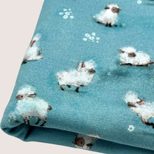 Load image into Gallery viewer, A piece of Cotton Jersey Fabric - Cute Sheep Aqua is shown in a close-up view, featuring a pattern of small, white sheep with black faces and legs, set against a blue background. This Oeko-Tex 100 certified dressmaking fabric from Once Upon A Fabric has the sheep scattered in various orientations across the fabric.
