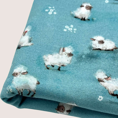 A piece of Cotton Jersey Fabric - Cute Sheep Aqua is shown in a close-up view, featuring a pattern of small, white sheep with black faces and legs, set against a blue background. This Oeko-Tex 100 certified dressmaking fabric from Once Upon A Fabric has the sheep scattered in various orientations across the fabric.