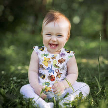 Load image into Gallery viewer, A baby with light hair sits on grass outdoors, smiling. The baby is wearing a sleeveless outfit made from Once Upon A Fabric&#39;s Organic GOTS Cotton Jersey - Happy Bugs and decorated with colorful cartoon insects and animals, including bees, ladybugs, frogs, and monkeys. Trees and greenery are blurred in the background.
