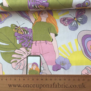 A fabric displaying a pattern with a woman in a green top and pink pants, surrounded by colorful butterflies, leaves, and abstract shapes. A hand is holding a smartphone capturing the design on this 97 cm REMNANT 100% Premium Cotton - Retro Summer material perfect for home furnishing from Once Upon A Fabric. A wooden ruler and website URL are visible at the bottom.