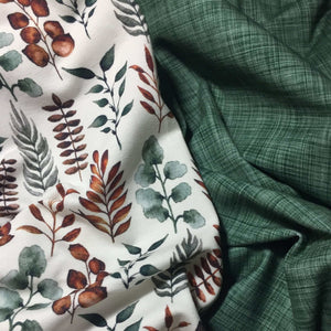 Close-up of two types of fabric, perfect for baby clothes. The left is white with a pattern of brown and green leaves on soft cotton jersey fabric. The right is green with a subtle woven texture. Both Cotton Jersey Fabric - Autumn Leaves by Once Upon A Fabric and Oeko-Tex 100 certified fabrics are slightly wrinkled and positioned next to each other.